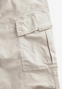 Pantaloni cargo color beige chiaro con una grande tasca a patta, cuciture visibili e un tessuto texture. Design funzionale con accesso laterale alle tasche.
