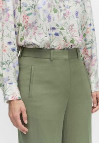 Lichtgroene op maat gemaakte broek met zijzakken, gecombineerd met een bloes in bloemenprint met pastelkleuren op een witte achtergrond.