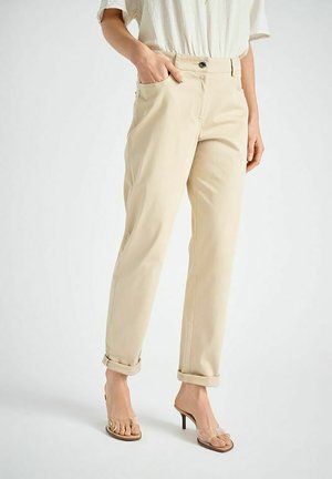 Vrouw draagt beige opgerolde broek, wit shirt met korte mouwen en hoge hakken met transparante bandjes, staat met één hand in de zak.