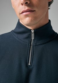 Navy strukturierter Viertel-Reißverschluss-Pullover mit hohem Kragen und silberner Reißverschlussbeschlägen, aus einem meshartigem Stoff.
