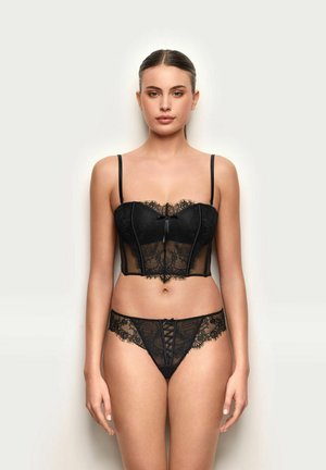 Donna in piedi davanti a uno sfondo semplice, indossa un bustino in pizzo nero con spalline sottili e slip coordinati in pizzo con dettaglio a fiocco sul davanti.