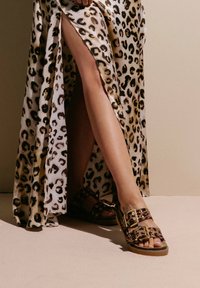 Maxikleid mit Leopardenmuster und hohem Schlitz, kombiniert mit braunen Sandalen mit Schnallen und Nietenakzenten. Glatter Stoff, kontrastierende Texturen.