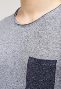 Zbliżenie na osobę w szarej, fakturowanej koszulce z ciemnoszarym kieszonką na piersi i widocznym dekoltem.