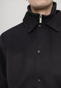 Black jacket with a collared neck, snap buttons, and a ribbed black inner collar. Smooth texture and minimalistic design.