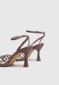 Sandales à talons en cuir brun métallique avec un design à pois transparent et des clous en argent sur la bride des orteils. Brides de cheville ajustables.
