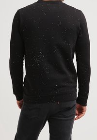 Persona che indossa una felpa nera con motivo a macchie bianche e jeans neri, vista di schiena su uno sfondo semplice.