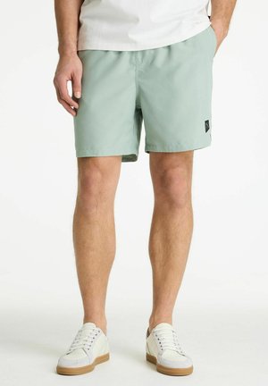 Férfi világoszöld rövidnadrágban és fehér tornacipőben, zsebében tartva a kezét, egyszerű fehér háttér előtt áll.