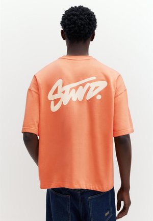 Persoon met kort krullend haar draagt een oversized perzikkleurig T-shirt met witte gestileerde tekst en donkerblauwe spijkerbroek, met de rug naar de kijker.