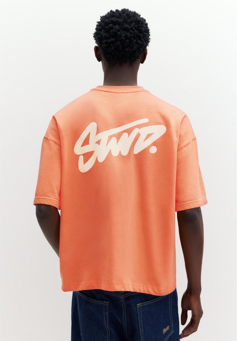 Personne aux cheveux courts et bouclés porte un t-shirt ample couleur pêche avec un texte blanc stylisé et un pantalon en denim bleu foncé, vue de dos.