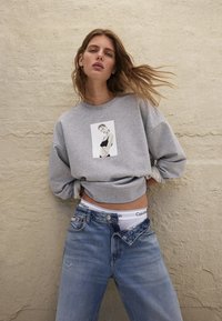 Donna con capelli sciolti indossa un felpa grigia con un ritratto in bianco e nero, jeans blu sbottonati e biancheria intima bianca Calvin Klein.