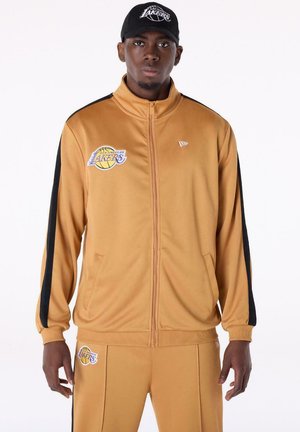 Mann in einem beigefarbenen Los Angeles Lakers-Trainingsanzug mit schwarzen Streifen und passender Kappe steht vor weißem Hintergrund.