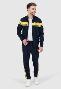 Marineblauer Trainingsanzug mit gelben Streifen, bestehend aus einer Reißverschlussjacke und Jogginghosen mit Kordelzug. Das Material wirkt glatt und beide Teile sind mit einem Logodetail versehen.