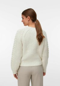 Vrouw met een bruine paardenstaart die een chunky witte gebreide trui en lichtbeige broek draagt, gezien vanuit de achterkant tegen een effen achtergrond.