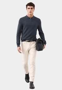 Marineblauw longsleeve polo met subtiele textuur, crème slim-fit broek en zwarte laarzen. Houdt een zwarte tas vast tegen een lichte achtergrond.