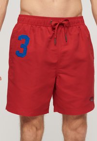 Shorts de bain rouges en tissu léger, avec une taille �élastique et un cordon de serrage, le numéro "3" en bleu sur la jambe gauche, avec peu de détails.
