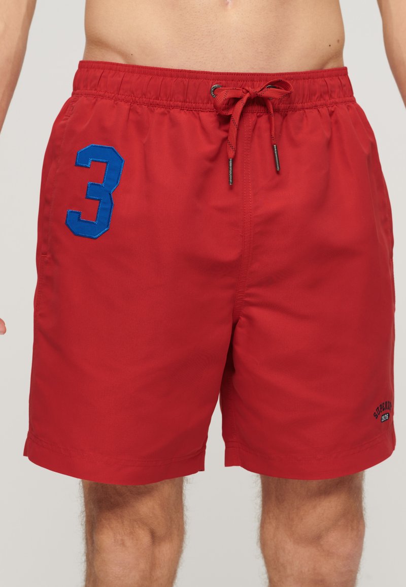 Shorts de bain rouges en tissu léger, avec une taille élastique et un cordon de serrage, le numéro "3" en bleu sur la jambe gauche, avec peu de détails.