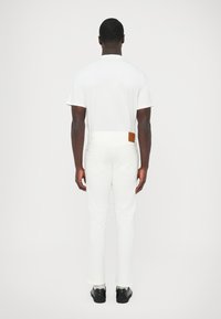 Λευκό εφαρμοστό τζιν με δερμάτινο ταμπελάκι στη μέση, σε συνδυασμό με απλό λευκό μπλουζάκι, φωτογραφημένο από πίσω. Φοράει μαύρα παπούτσια.