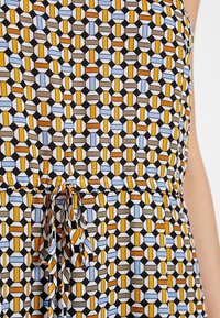 Sukienka z geometrycznym wzorem z przeplatającymi się heksagonami w kolorach żółtym, niebieskim, brązowym i czarnym, z wiązanym paskiem w talii.