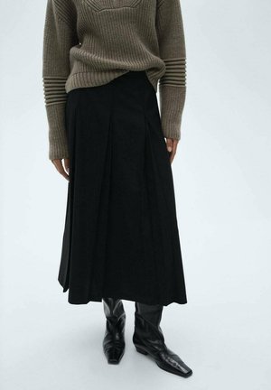 Person wears a ribbed brown sweater, black pleated midi skirt, and black pointed square-toe mid-heel boots, standing on a plain light background.
