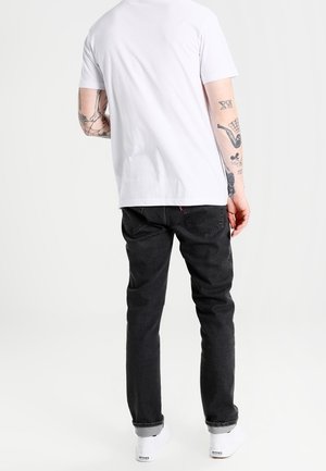 Camiseta blanca de manga corta, vaqueros negros con un ligero elastano, ajuste informal, tatuajes visibles en el brazo y zapatillas blancas.