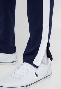 Marina byxor med vita sidosträck, med en avsmalnande design. Matchas med vita sneakers som visar en logotyp och kontrasterande blå detaljer.