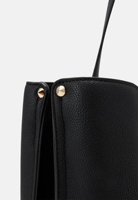 Czarna skórzana torebka o teksturowanym wykończeniu, z złotymi akcentami hardware i eleganckim designem. Dwa okrągłe ćwieki po bokach.