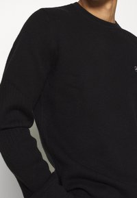 Maglione nero a maglia con un design a coste testurizzato, scollatura rotonda e maniche lunghe aderenti. Presenta un discreto dettaglio logo sul petto.