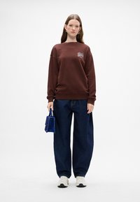 Sudadera marrón con un logo en el pecho, combinada con unos vaqueros oscuros holgados y zapatillas blancas. El modelo sostiene un pequeño bolso azul.