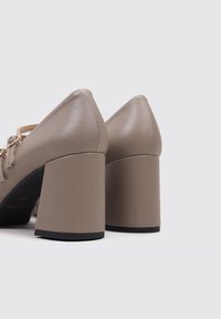 Chaussures à talons hauts beige avec une finition en cuir brillant, talons carrés, et un design arrière pointu. Des accents métalliques sont présents.