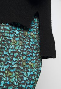 Svart tröja med mjuk textur, bärs över en flerfärgad mönstrad klänning med gröna och blå abstrakta former på en mörk bakgrund.