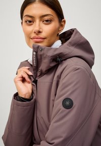 Chaqueta de cremallera en color malva claro con cuello alto, capucha ajustable y parche con logo en la manga izquierda. Fabricada con material liso y resistente al agua.