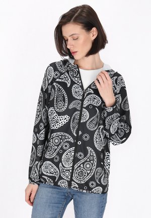 Giacca nera con intricati motivi paisley bianchi, chiusura con zip e cappuccio. Realizzata in materiale morbido, abbinata a jeans azzurri chiari.