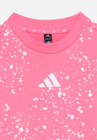 Felpa rosa con colletto a coste, caratterizzata da un motivo a macchie bianche e un logo Adidas bianco sul petto. Tessuto di cotone liscio.