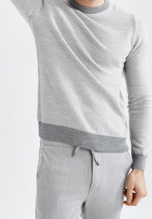 Mann trägt hellgrauen Fischgrät-Pullover und hellgraue Hose mit Kordelzug, hebt den linken Arm.