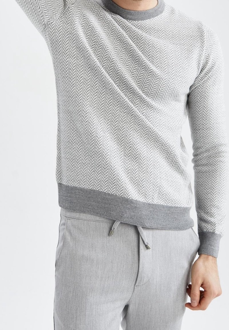 Homme portant un pull gris clair à motif chevrons et un pantalon gris clair avec cordon, levant le bras gauche.
