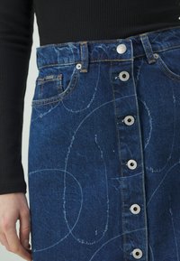 Jeanskjol med blå tvätt, utrustad med silverknappar, två framfickor och vita abstrakta mönster på tyget.