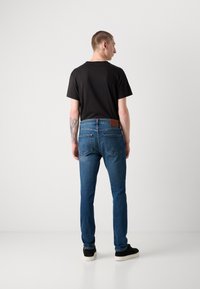 Blaue Jeans aus Denim im Slim-Fit-Schnitt, mit Gesäßtaschen und einem braunen Lederlabel am Bund. Kombiniert mit einem schwarzen kurzärmeligen Hemd.