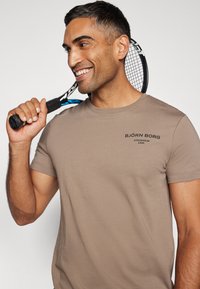Brun kortermet T-skjorte med trykket logo, rund hals, mykt bomullsmateriale, båret av en person som holder et tennisrack.