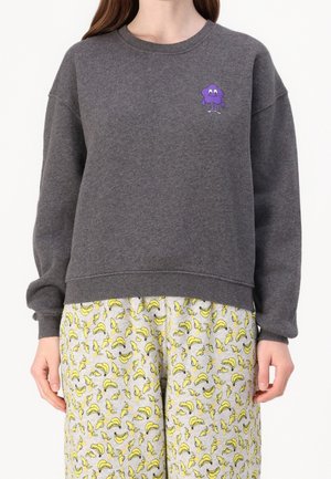 Henkilö, jolla on tummaharmaa collegepaita, jossa on pieni violetti sarjakuvahahmo rinnan kohdalla, ja vaaleat housut, joissa on keltainen banaanikuviointi.