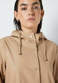 Veste imperméable beige avec une capuche, des boutons-pression et des cordons de serrage. Fabriquée en matériau lisse et léger, avec un design minimaliste.