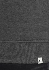 Donkergrijze sweatshirt met een geribbelde zoom. Heeft een klein wit logotag aan de onderkant links. Zacht, textuurstof.
