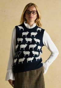 Tumes sinine kootud vest, millel on valgete lambade muster, ümar kaelus ja ribaga äär. Kantud valge pluusi peal, millel on volangidega varrukad.