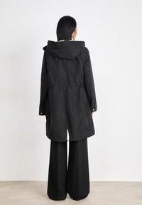 Zwarte waterdichte parka met capuchon, taillen accentueerd en knopen. Lange mouwen en rechte zoom, gecombineerd met wijde zwarte broeken.