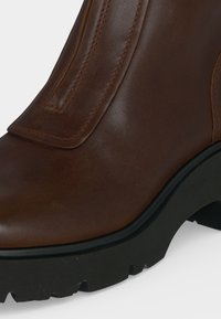Gros plan sur une botte en cuir marron avec une semelle épaisse en caoutchouc noir et des coutures apparentes, montrant les détails à l'avant et sur le côté.