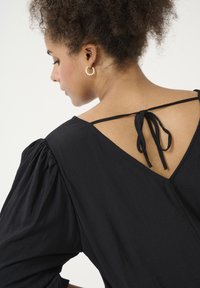 Blouse noire avec manches bouffantes, décolleté en V et un accent noué à l'arrière. Texture lisse et tissu léger.