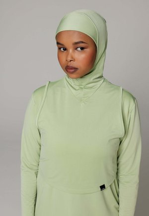 Vrouw draagt een lichtgroene aangesloten hijab en bijpassende longsleeve, kijkt opzij tegen een effen grijze achtergrond.