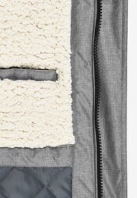 Gros plan sur une veste montrant un tissu gris, une fermeture éclair noire, une doublure en polaire blanche et une petite poche bordée de tissu.