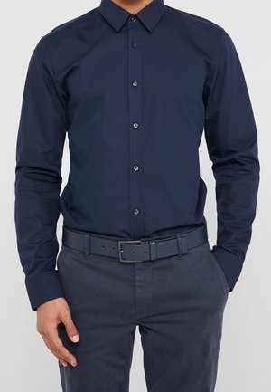 Man die een nauwsluitend marineblauw overhemd met knopen draagt, ingestopt in bijpassende marineblauwe broek met een marineblauwe riem, handen ontspannen langs het lichaam.