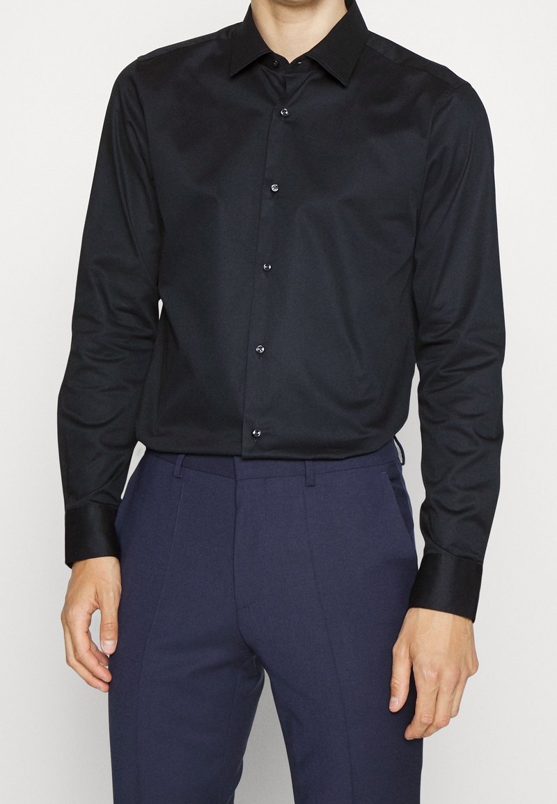 Homme portant une chemise noire cintrée boutonnée, rentrée dans un pantalon bleu foncé sur mesure, debout devant un fond clair uni.