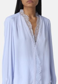 Blusa blu chiaro realizzata in un morbido tessuto, con bordatura in pizzo lungo il colletto e maniche arricciate per una silhouette fluida.